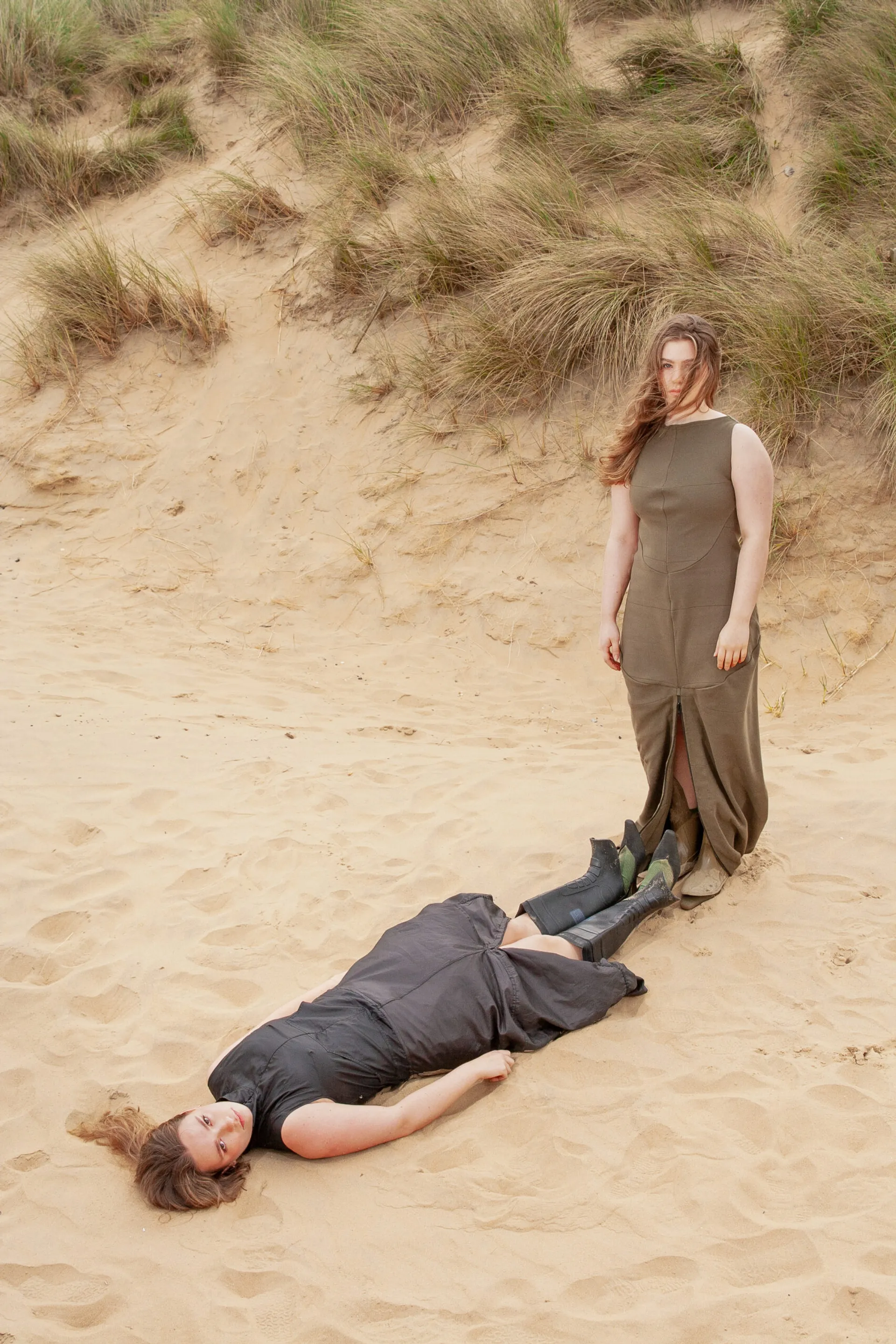 transformative pac-away black nylon dress worn long by model laying down in the sand dunes with wellie boot heels. model standing up with feet placed at feet of model laying down wears a similar dress in a jersey rib. both models reflect each other in their pose but also in appearance as they are both white women with long light brown hair.